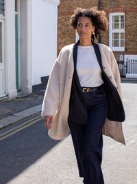 Woman walking on a street wearing a beige coat, white shirt, and dark pants.