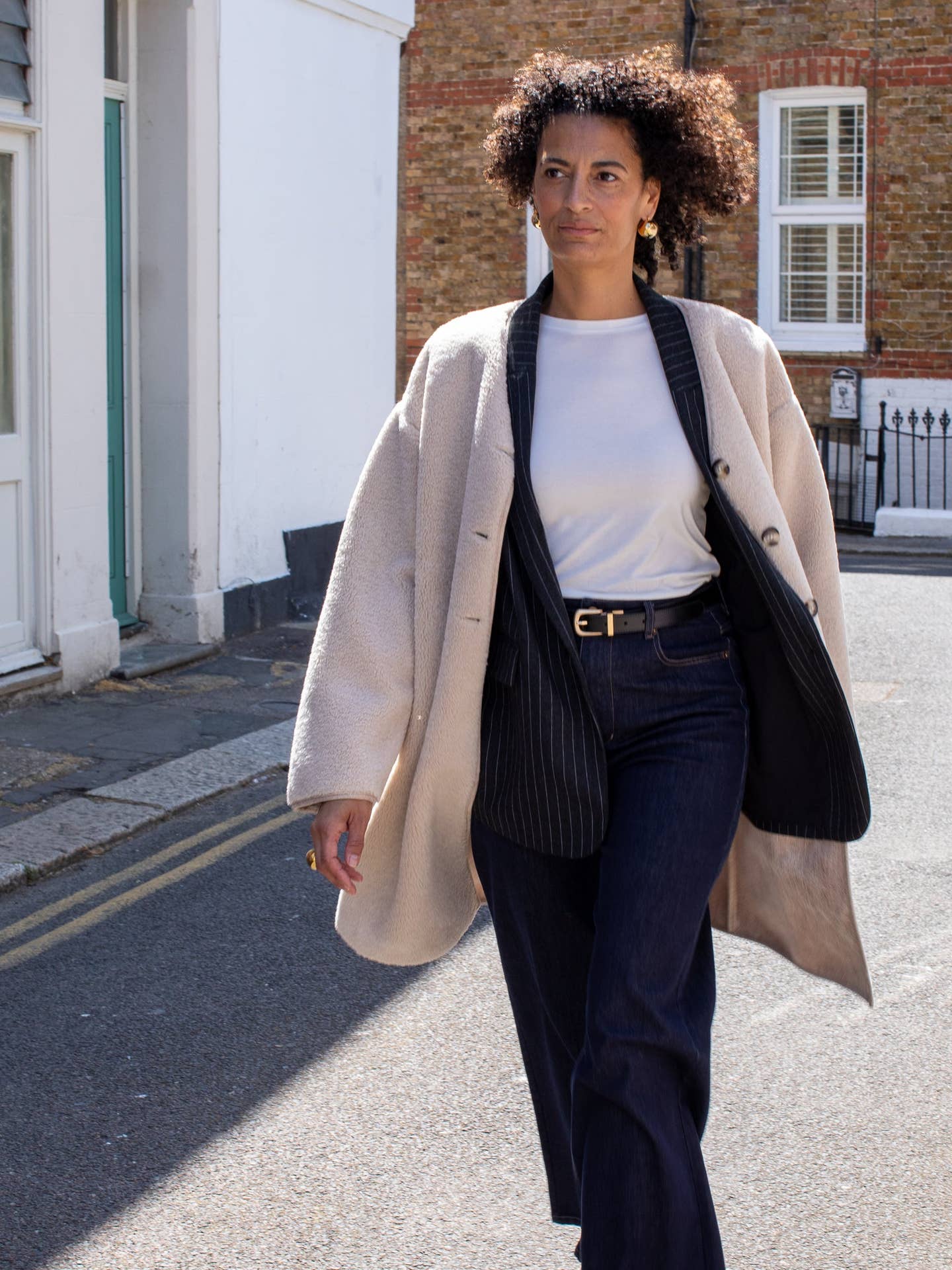 Woman walking on a street wearing a beige coat, white shirt, and dark pants.