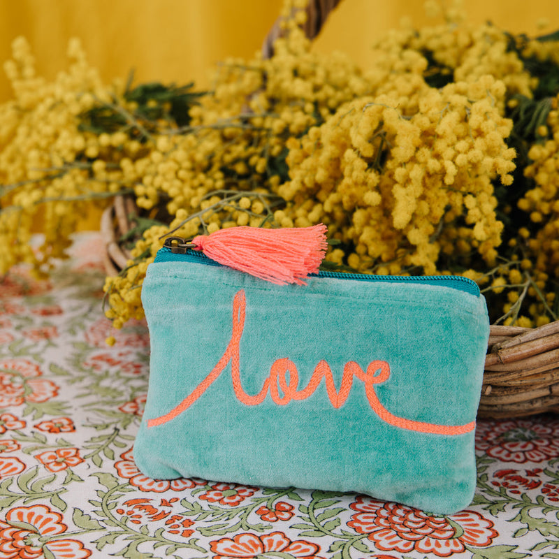 Turquoise pouch with 'love' embroidery and pink tassel on a floral tablecloth with yellow flowers in the background.