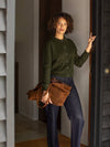 Woman in a green sweater and blue jeans holding a brown leather bag, standing in a doorway.