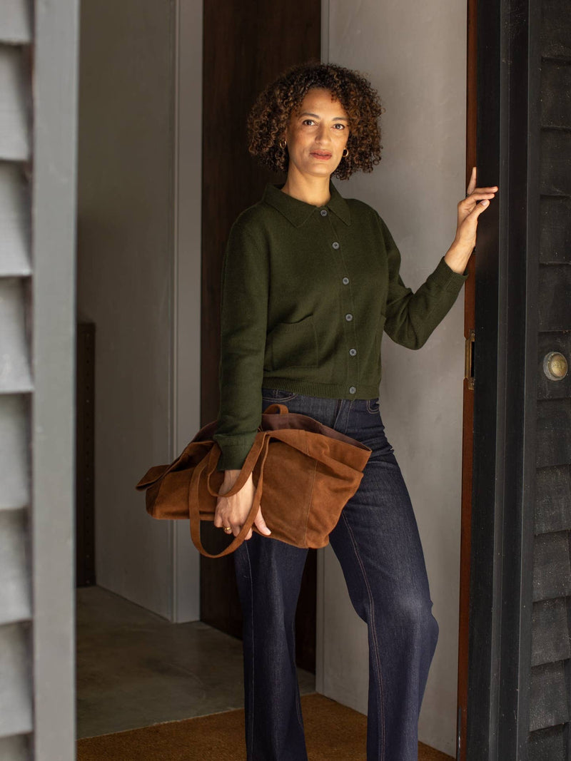 Woman in a green sweater and blue jeans holding a brown leather bag, standing in a doorway.