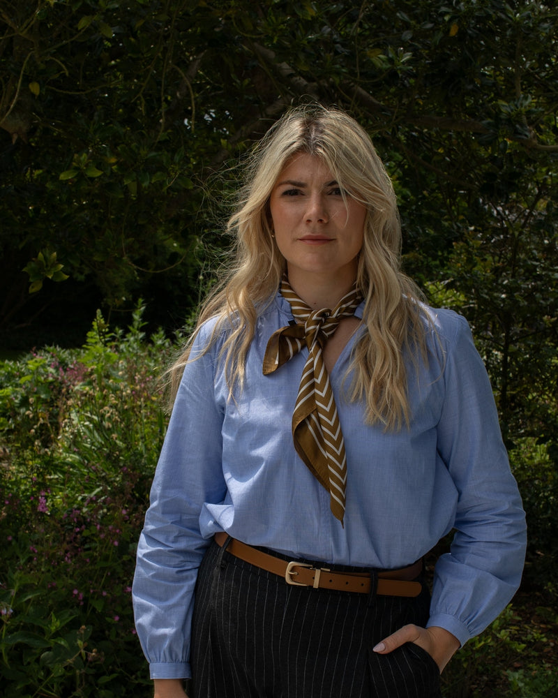 Woman wearing a blue shirt with a brown scarf and black pants outdoors.