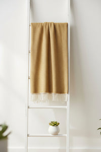 A rectangular gold woven throw blanket with a subtle herringbone texture and cream fringe along the bottom edge, shown flat against a solid white background.