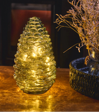A pinecone-shaped glass candle holder with warm yellow LED lights inside, set on a reflective metallic surface next to dried flowers in a dark, moody interior setting.