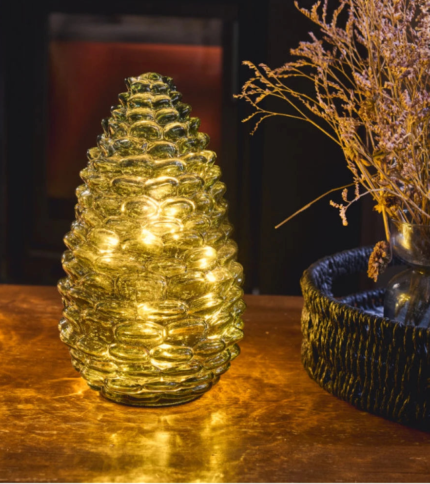 A pinecone-shaped glass candle holder with warm yellow LED lights inside, set on a reflective metallic surface next to dried flowers in a dark, moody interior setting.