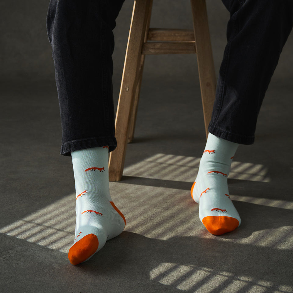 Person wearing light blue socks with orange tips and fox patterns, standing on a dark floor.
