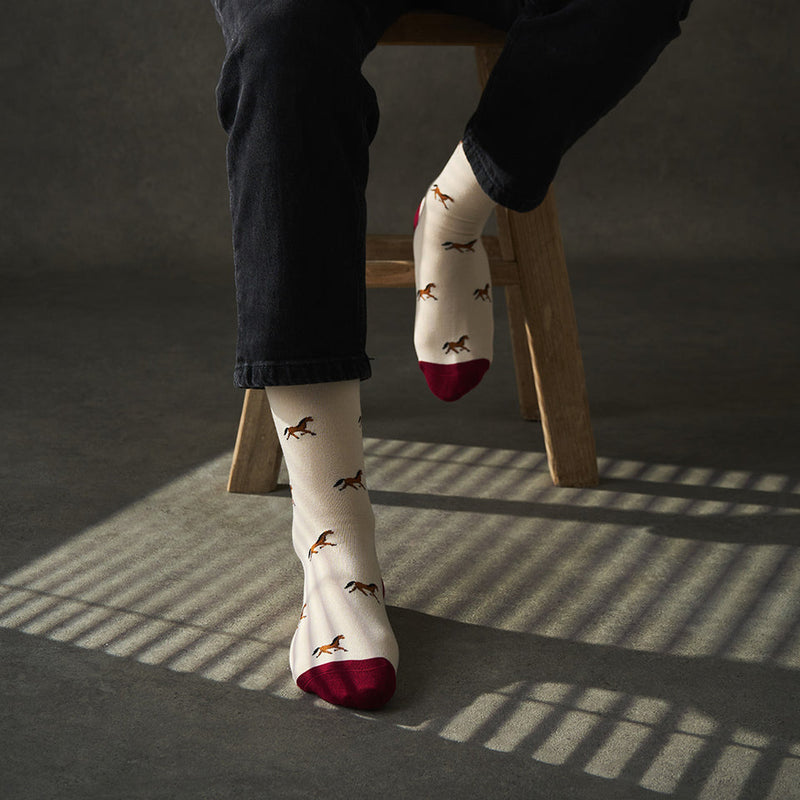 Person wearing socks with horse pattern and red toes, sitting on a wooden stool.