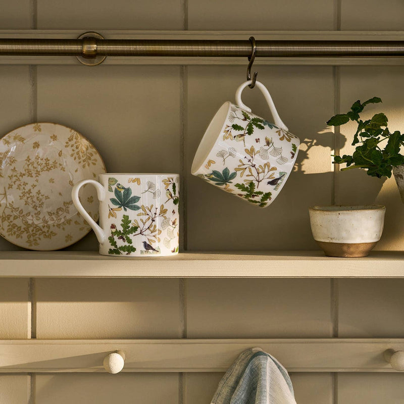 Two floral mugs hanging on a hook with a plate and plant in the background.