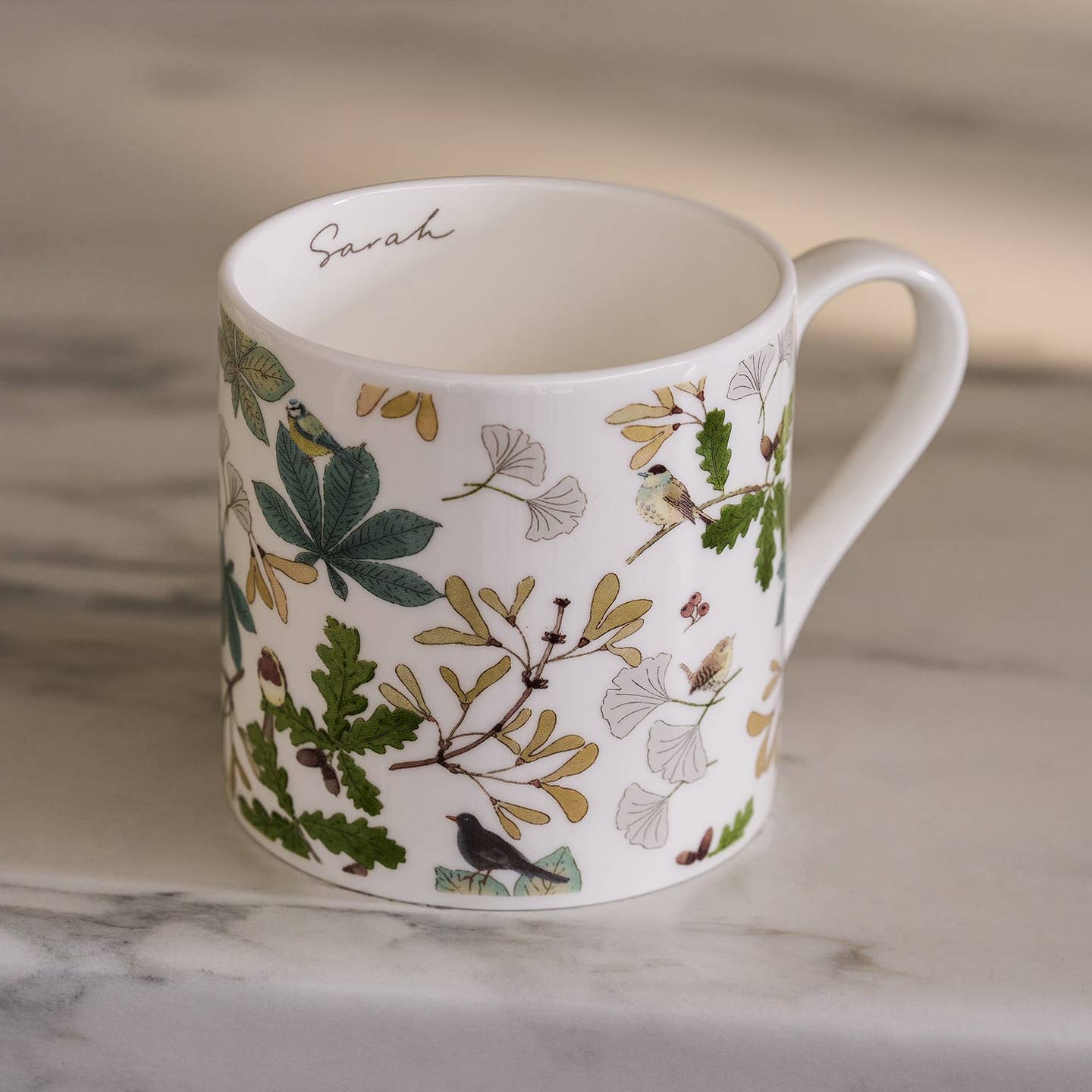 White mug with nature-themed design featuring leaves and birds on a marble surface.
