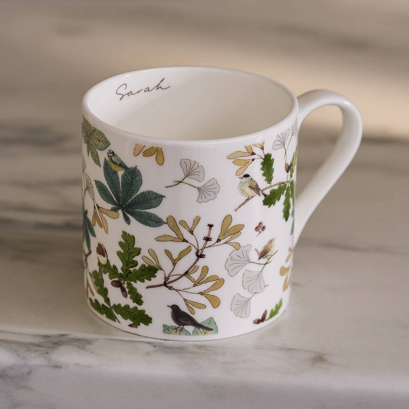 White mug with nature-themed design featuring leaves and birds on a marble surface.