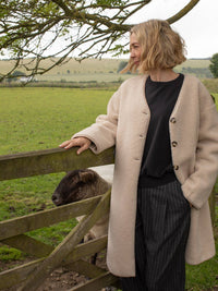 Woman in a beige coat standing next to a wooden fence with a sheep in a field.