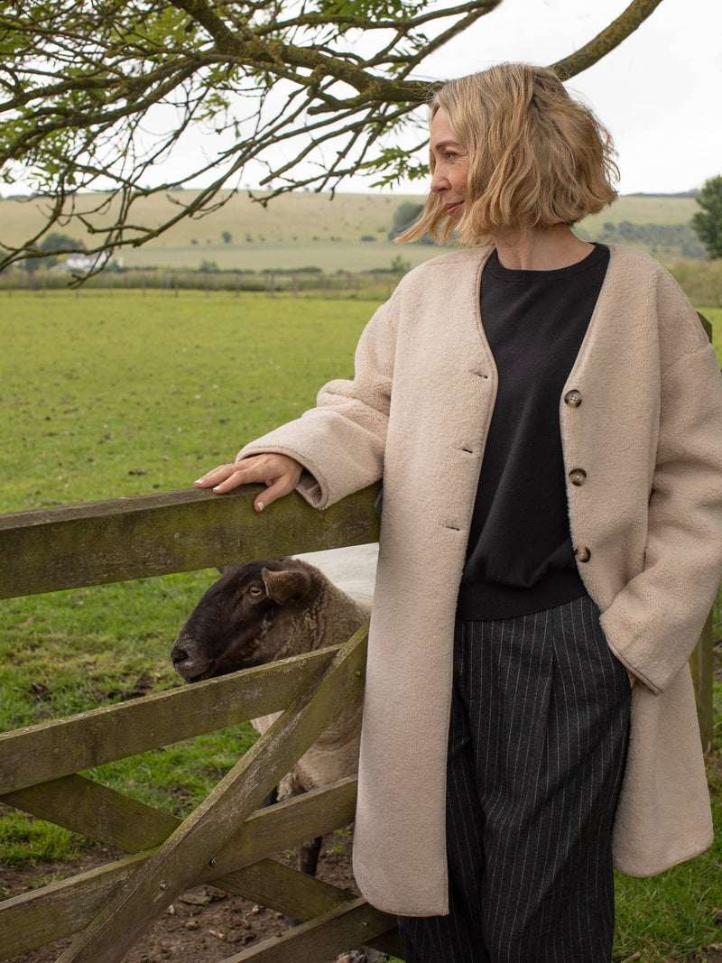 Woman in a beige coat standing next to a wooden fence with a sheep in a field.