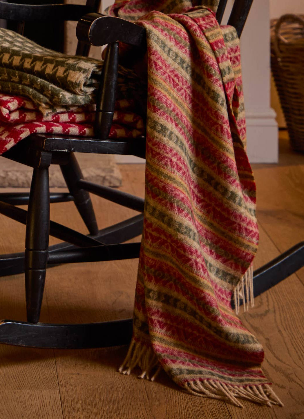 A patterned woven throw blanket draped over a dark wooden rocking chair in a cosy indoor room with wood floors and a basket in the background.
