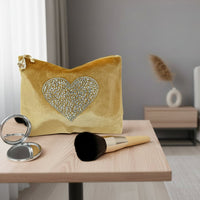 Gold clutch bag with a heart-shaped gemstone design, makeup brush, and compact mirror on a white background
