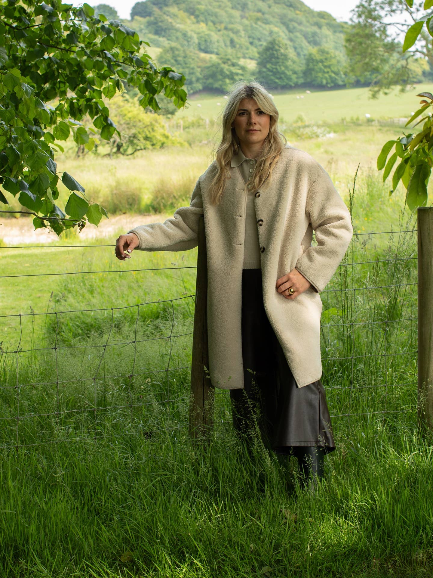 Woman in a beige coat standing in a grassy field with trees and hills in the background