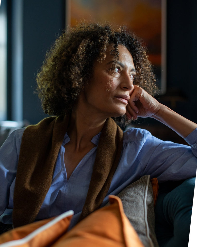 Woman sitting on a couch with a thoughtful expression in a dimly lit room.