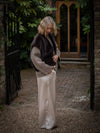 Woman in a brown sweater and white pants standing in an outdoor setting with a door and plants in the background.