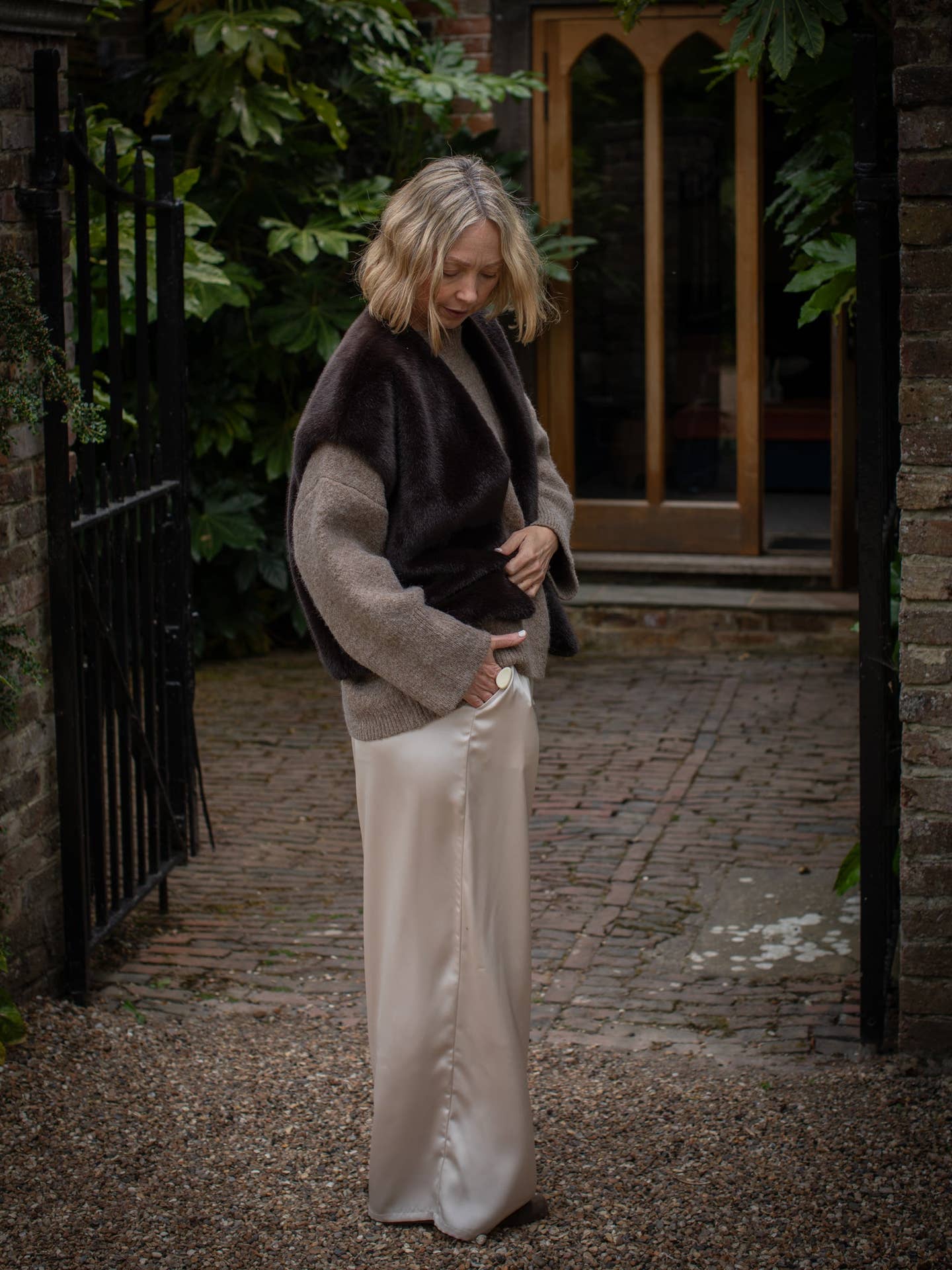 Woman in a brown sweater and white pants standing in an outdoor setting with a door and plants in the background.