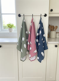 Set of three patterned sheep tea towels hanging on a rack with a kitchen background.