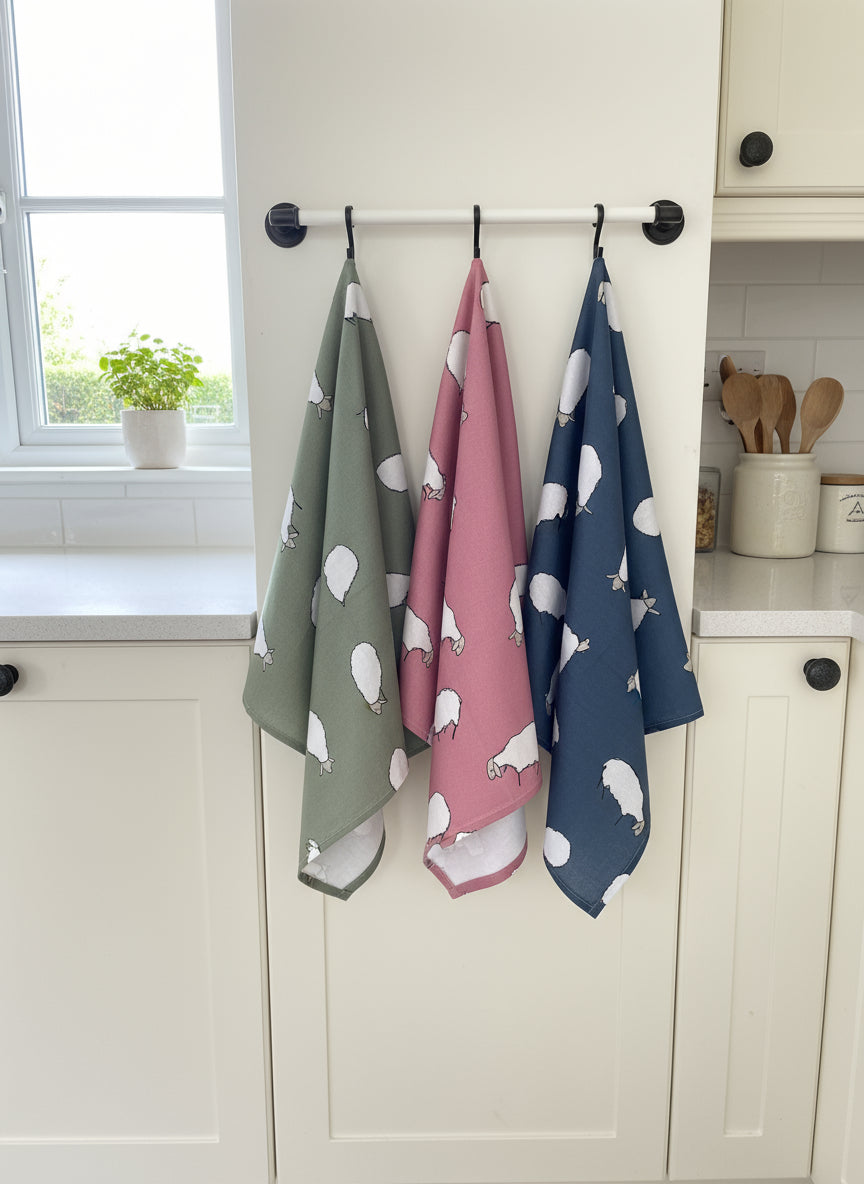 Set of three patterned sheep tea towels hanging on a rack with a kitchen background.